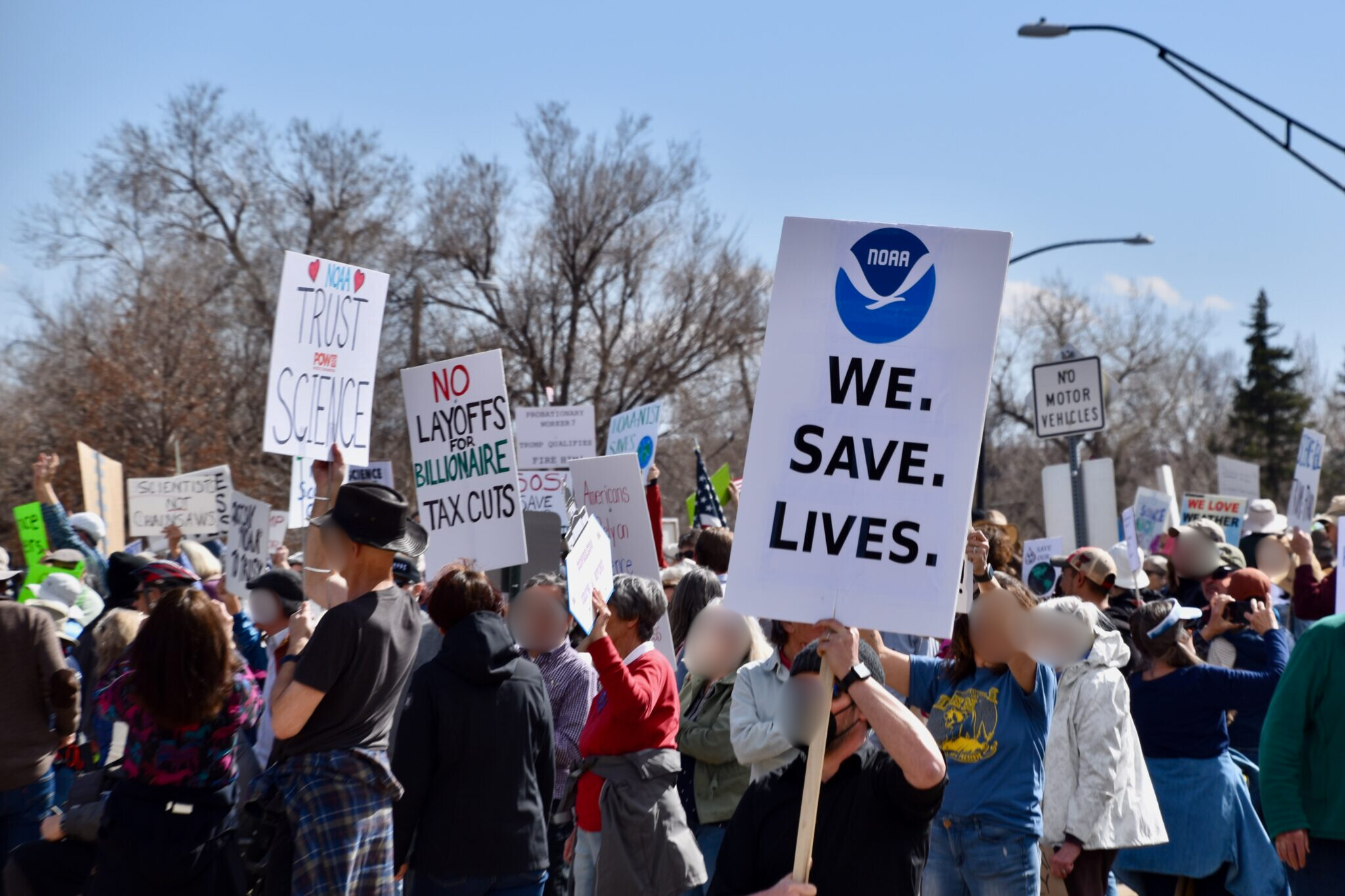 People holding up protest signs. One has a large print of the NOAA logo and says 'we save lives'.